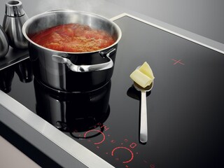 Uma colher com manteiga pousada em segurança numa placa fria ao lado de um tacho com molho de tomate em ebulição