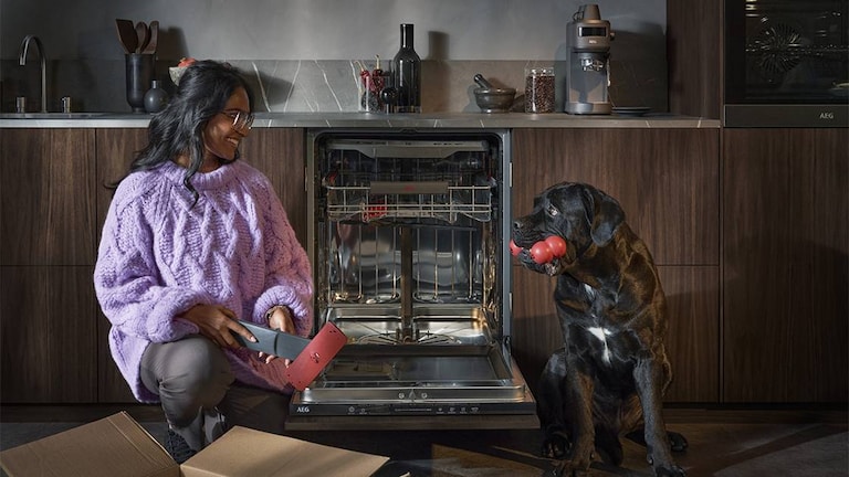 Mulher a desembalar peças de substituição para máquina de lavar loiça, enquanto sorri para um cão preto com um brinquedo na boca